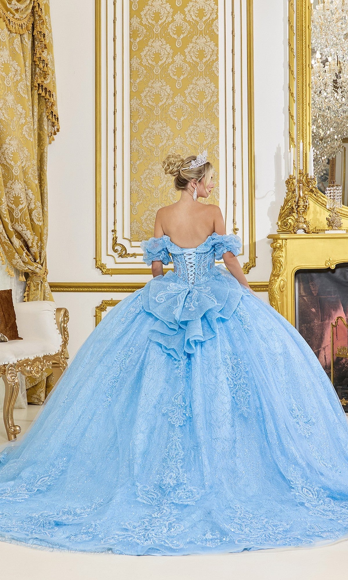 A woman wearing the Quinceanera Dress 5100 by Dancing Queen, an off-the-shoulder blue ballgown, stands with her back to the camera in an ornate room with gold decor and a grand fireplace.